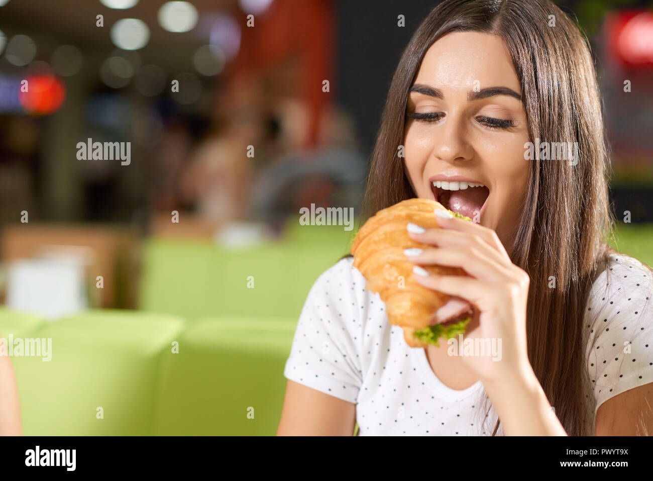 Hungry Female Client Sitting In Cafe And Biting Delicious Croissant With Greens And Ham