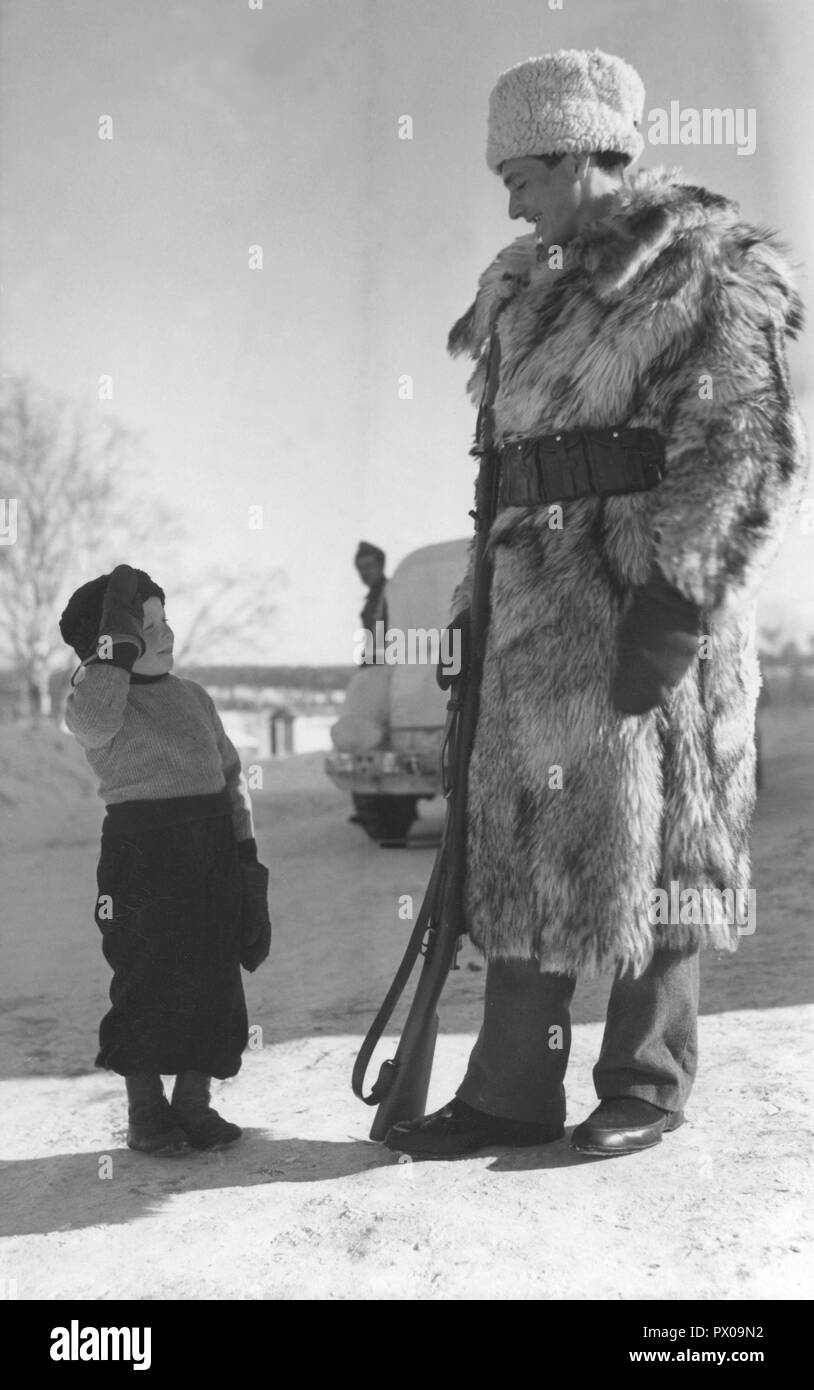 Swedish army during WW2. A soldier is standing on guard on a cold winters  day and is wearing a fur coat. A small boy has stopped to salute the  soldier. Sweden 1940s