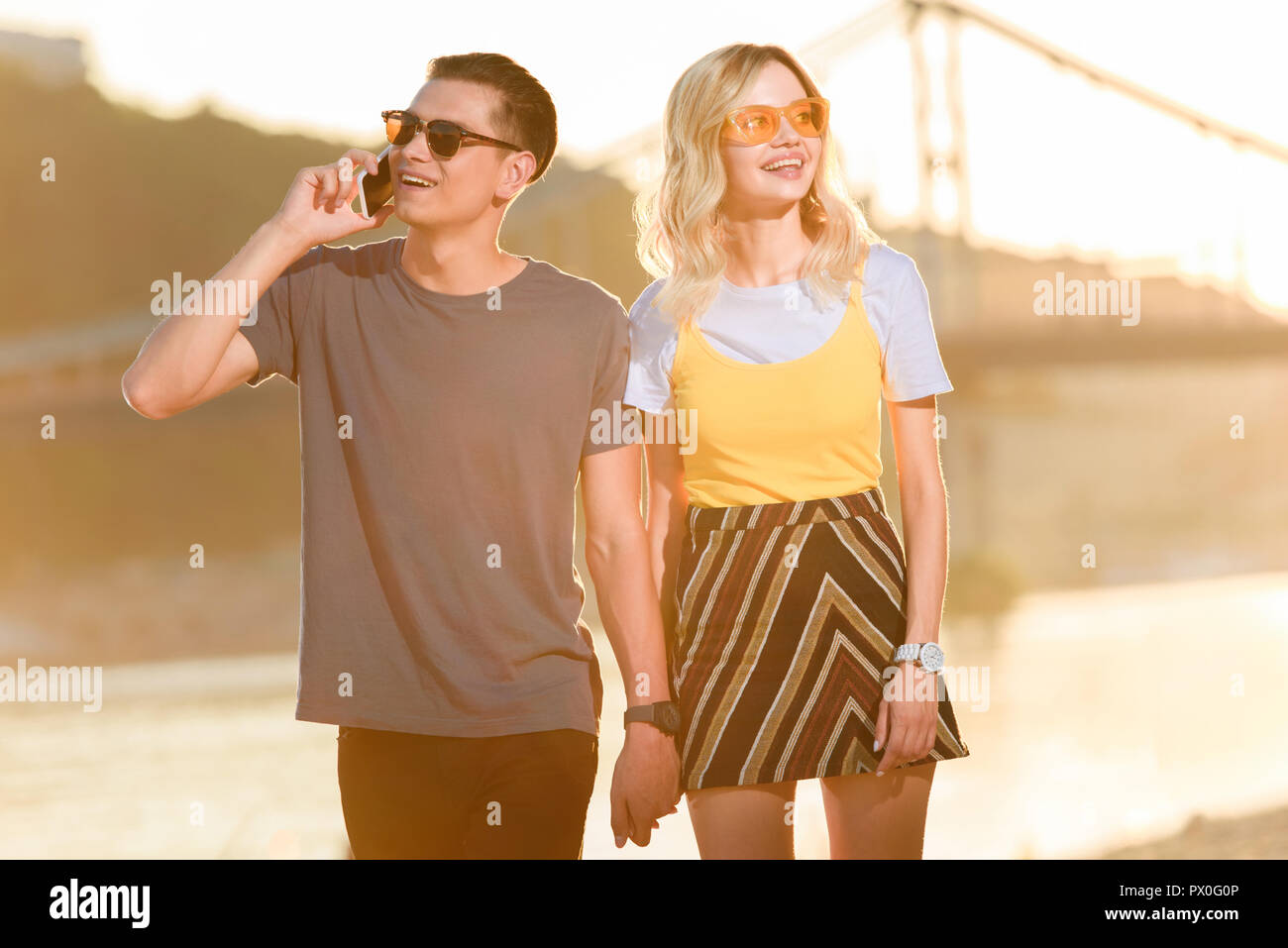 young couple walking on river beach in evening, boyfriend talking by smartphone Stock Photo