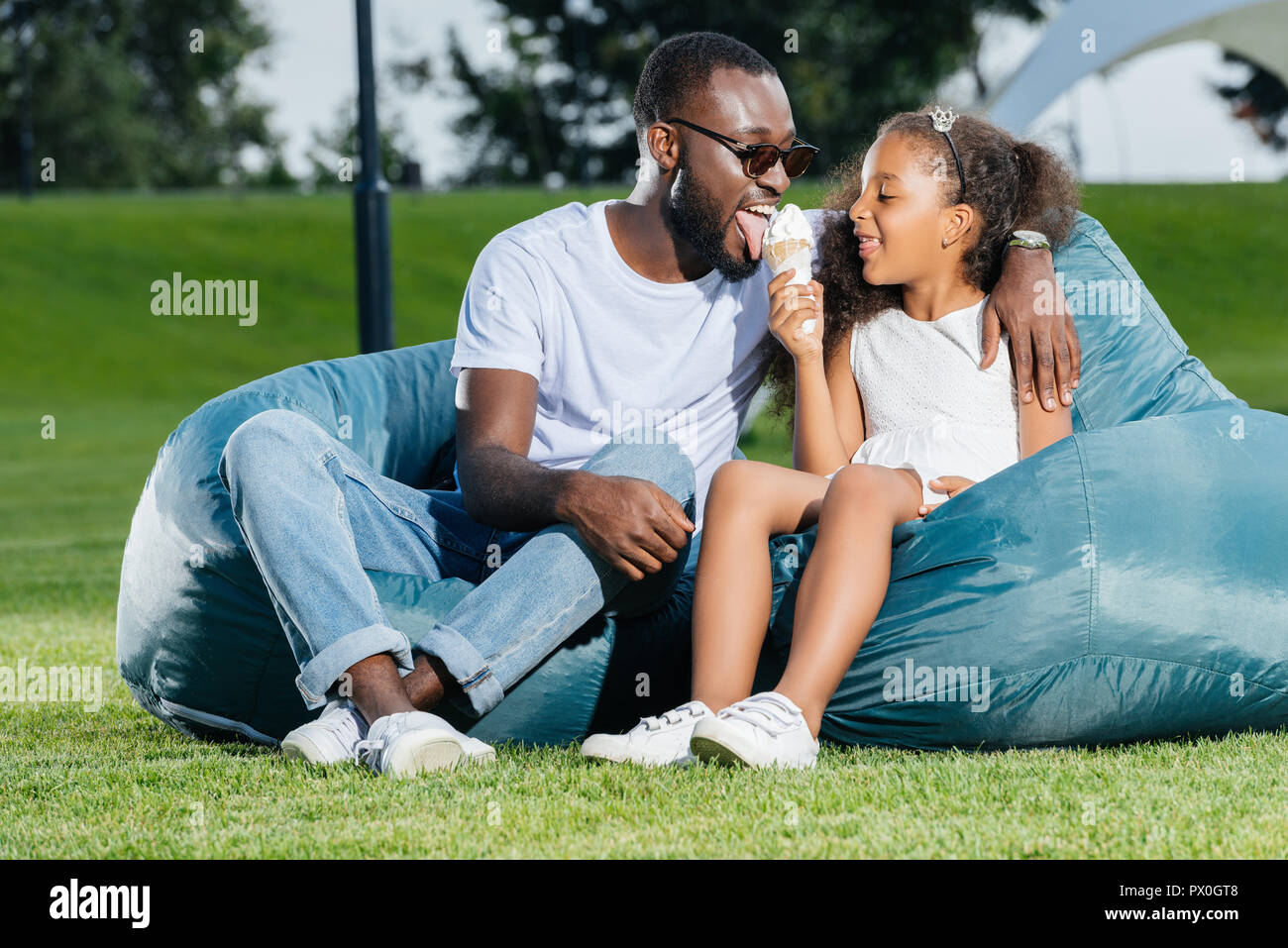 African American Father Licking Ice Cream And Sitting On Beanbag Chairs With Daughter In Park Stock Photo Alamy