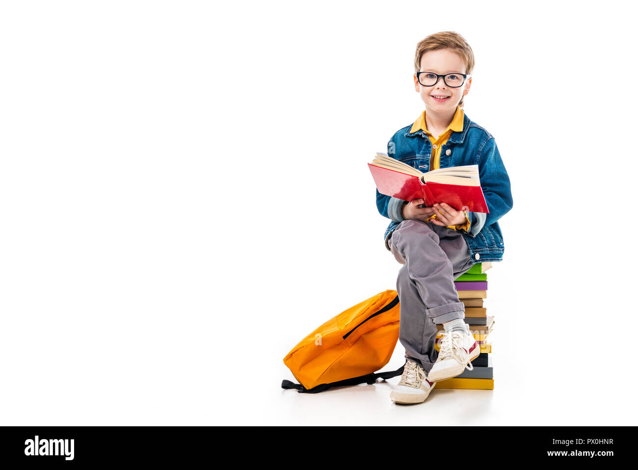 cheerful schoolboy reading book and sitting on pile of books with backpack, isolated on white Stock Photo