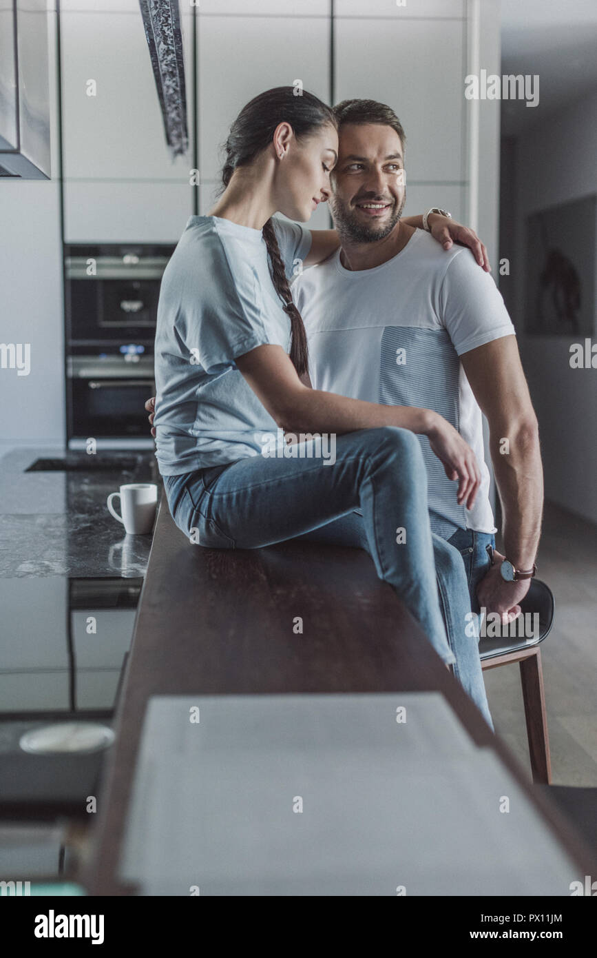 side view of attractive woman sitting on kitchen counter and embracing ...