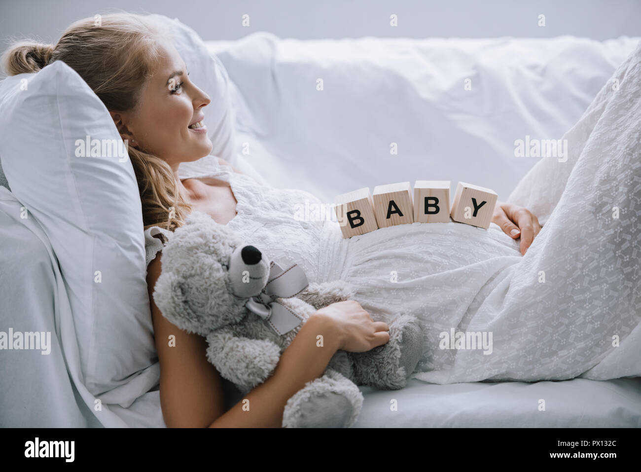 side view of smiling pregnant woman in white nightie with teddy bear and wooden blocks with baby lettering on belly resting on sofa Stock Photo
