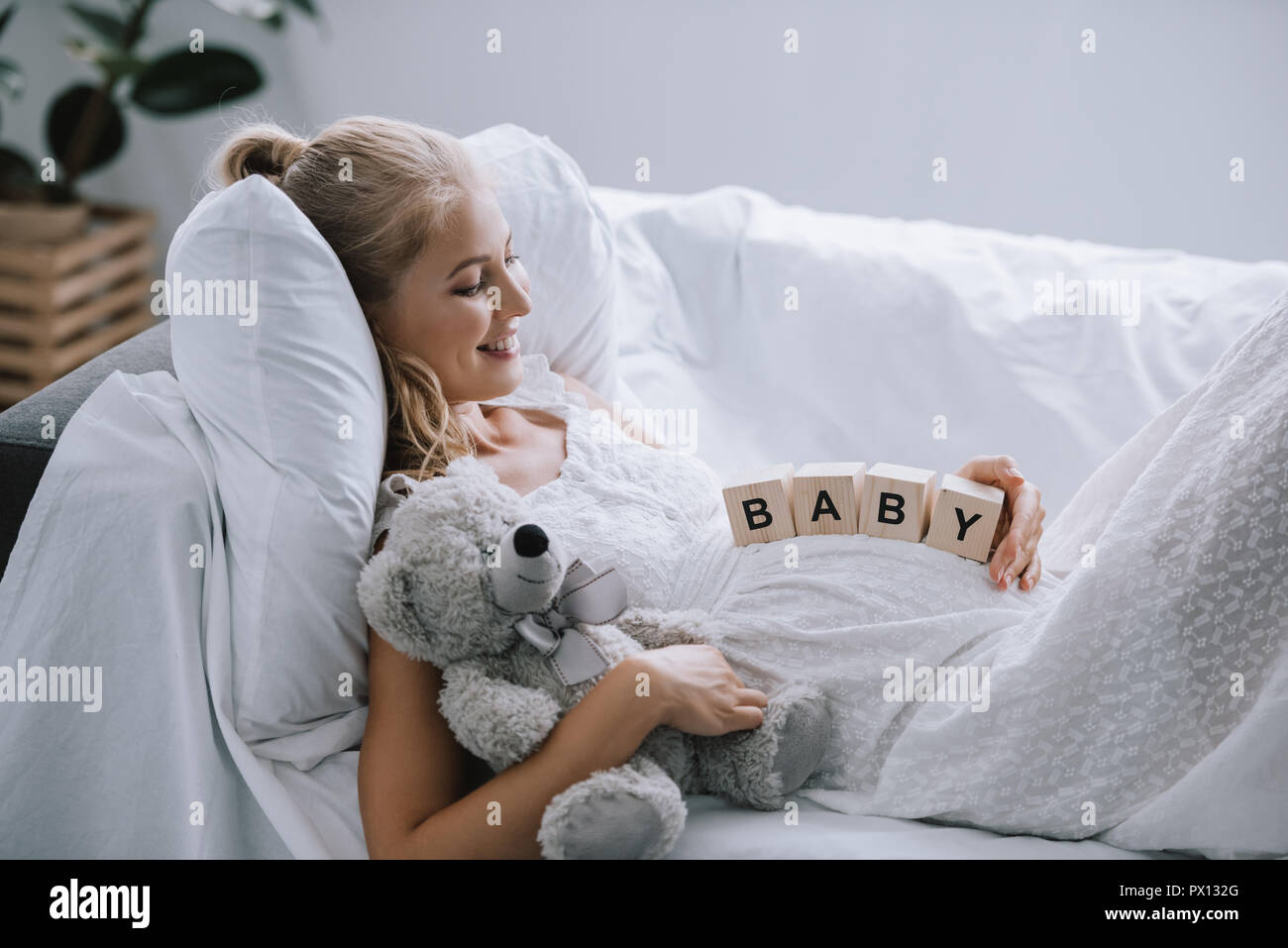 side view of smiling pregnant woman in white nightie with teddy bear and wooden blocks with baby lettering on belly resting on sofa Stock Photo