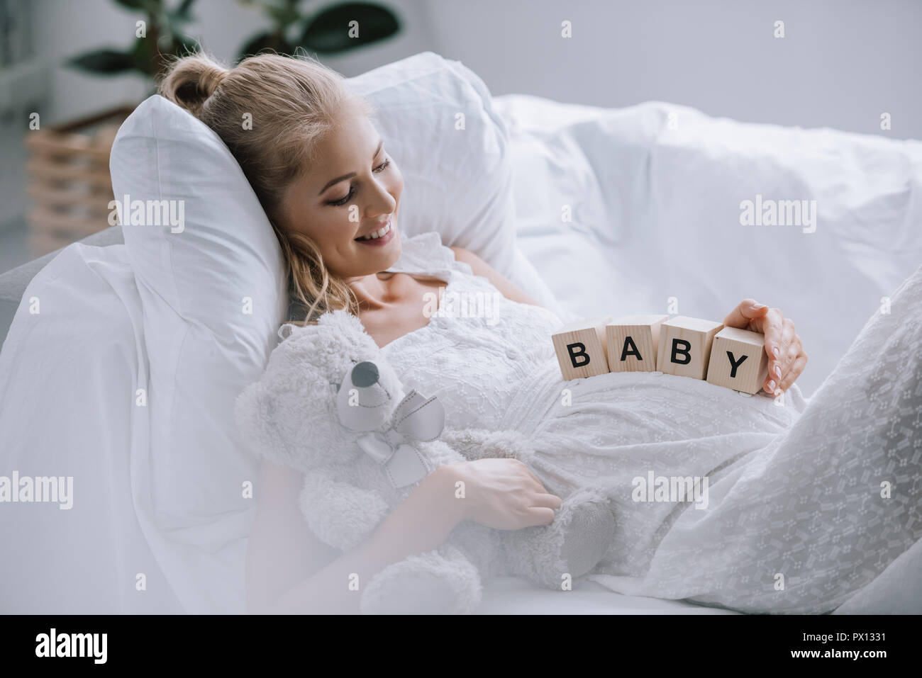 side view of smiling pregnant woman in white nightie with teddy bear and wooden blocks with baby lettering on belly resting on sofa Stock Photo