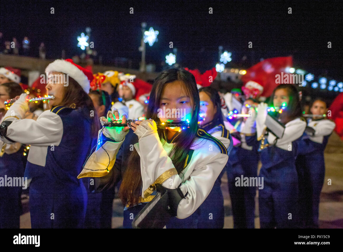 An Asian American uniformed high school band flutist plays with fellow band  members at a Christmas celebration in Huntington Beach, CA, as illuminated  Christmas stars are lit on the pier in the