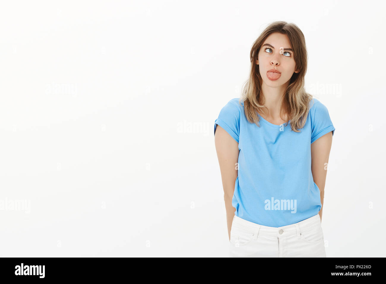 Portrait of positive childish and emotive girlfriend in blue t-shirt, holding hands behind back, squinting and sticking out tongue, making faces or aping while having great time with little brother Stock Photo