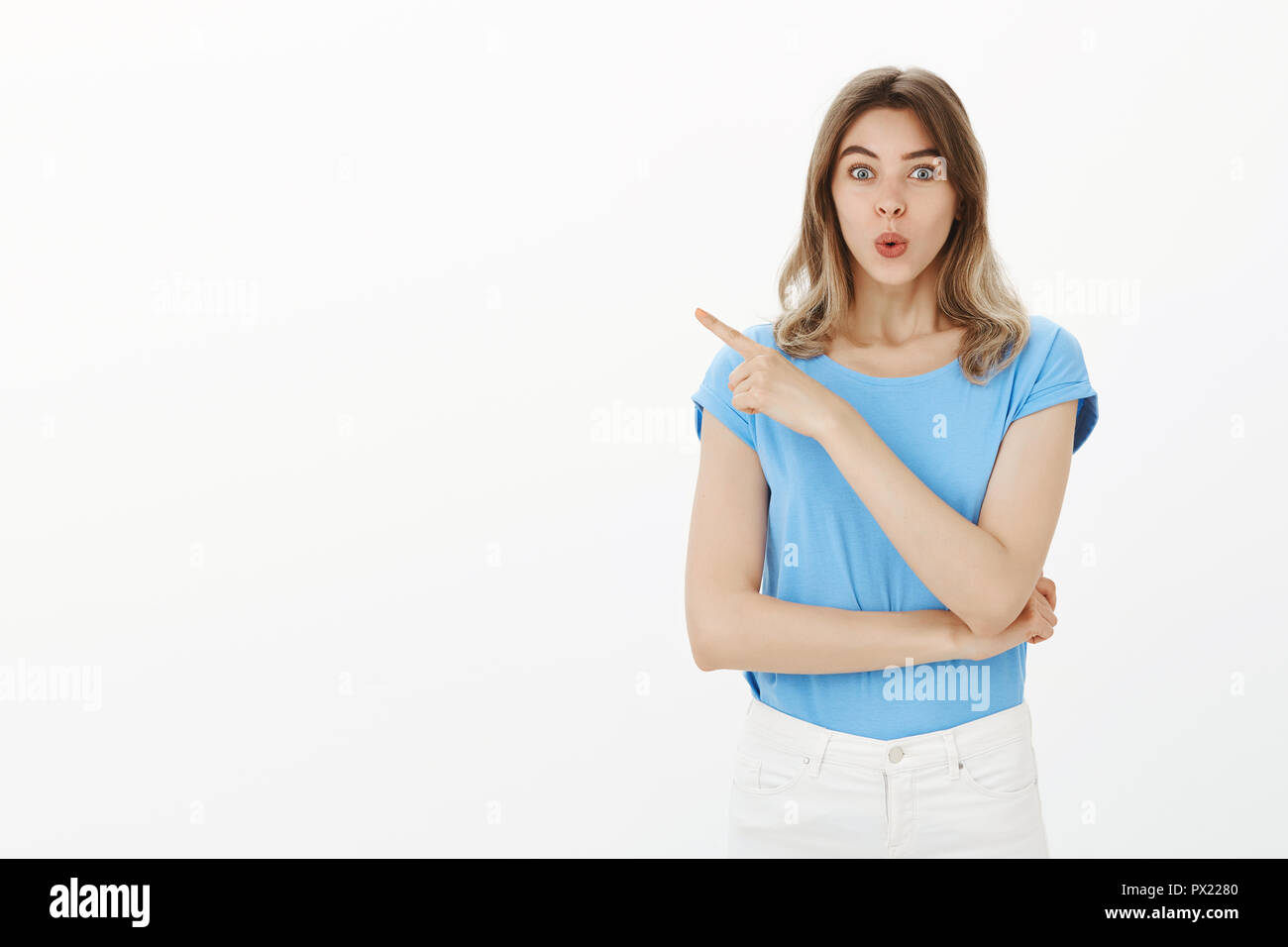 Wow, cannot wait to see this new film. Portrait of feminine good-looking woman in blue t-shirt, folding lips and pointing at upper left corner while discussing something with temptation over grey wall Stock Photo