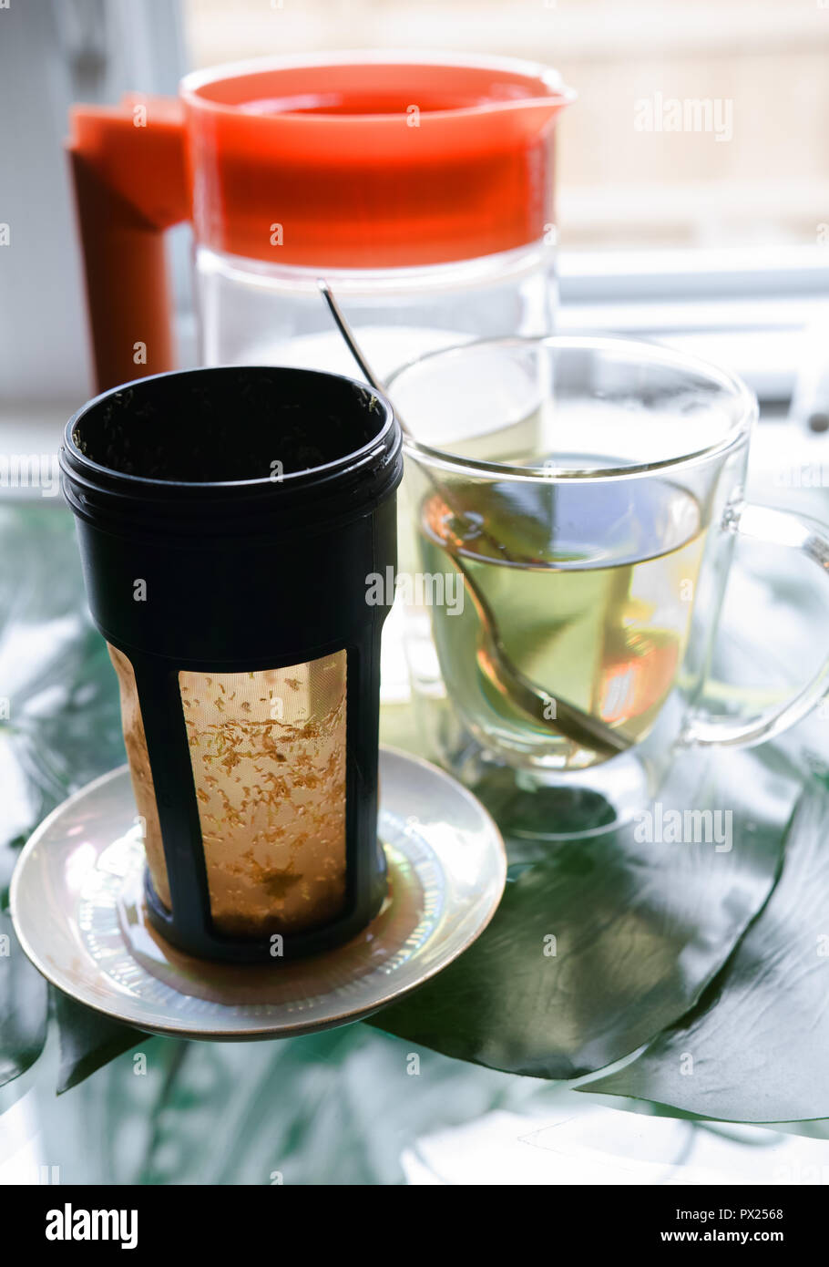 In the foreground there is the tea filter, then we have the transparent glass cup with pale tea in it and the spoon. Behind, we have a transparent pot Stock Photo