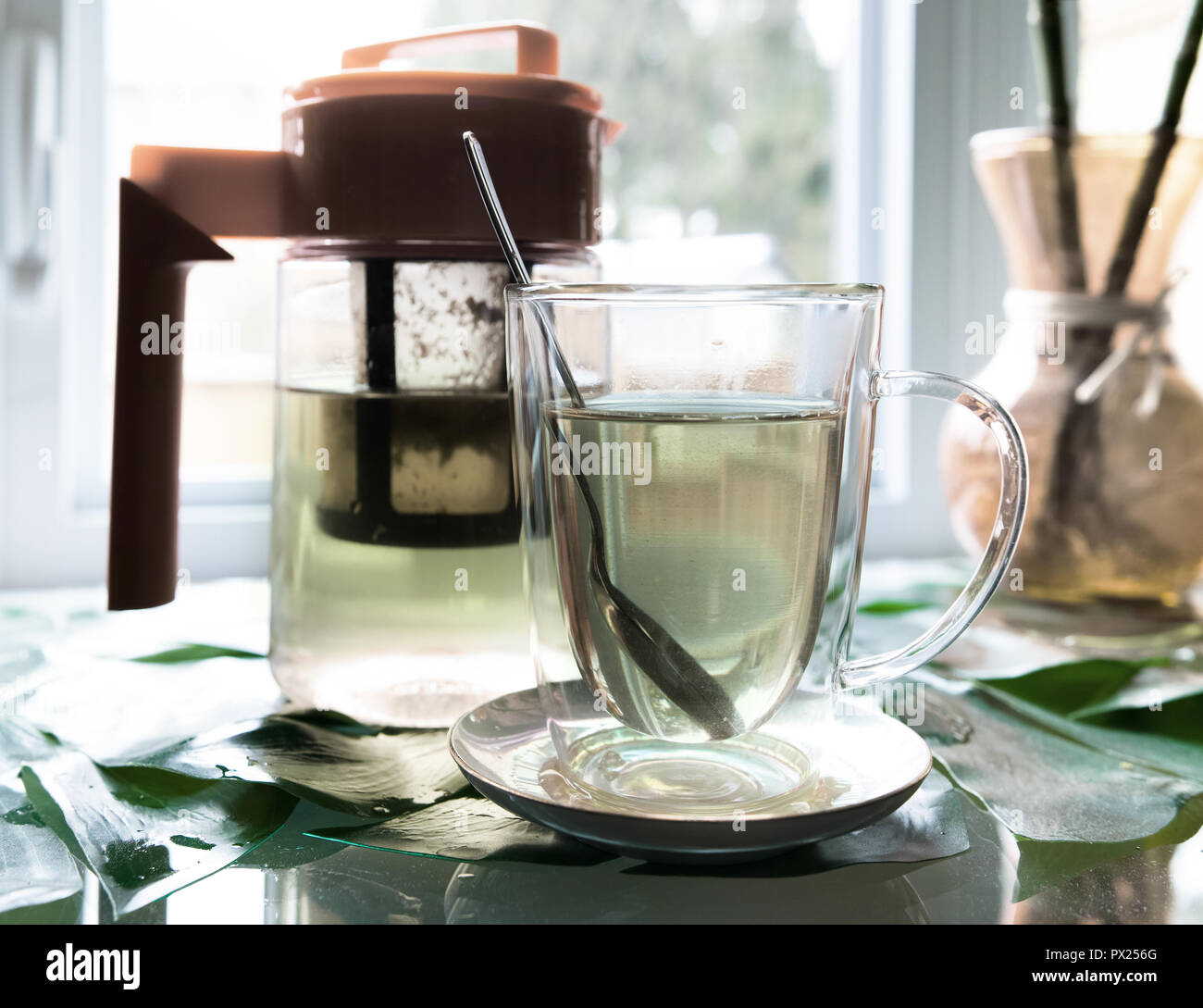 In front of the window there is a transparent pot of tea as well as a cup of tea with the spoon in it. Stock Photo