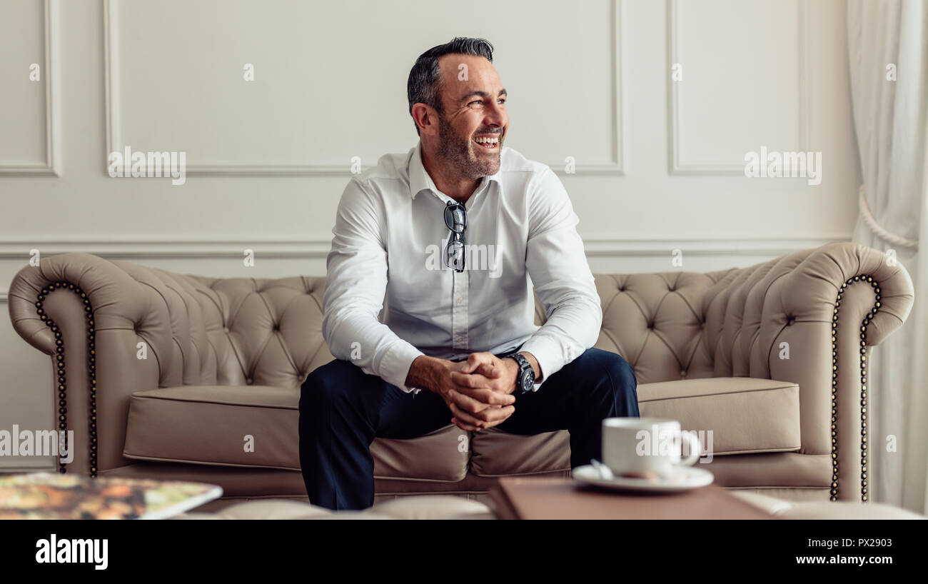 Portrait of cheerful businessman sitting on sofa in hotel room and looking away laughing. CEO staying in luxurious hotel room on business trip. Stock Photo