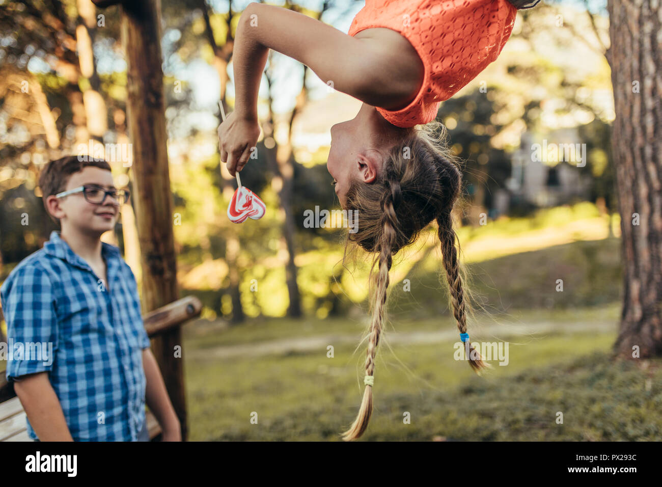 Girl hanging upside down in a park holding a heart shaped candy lollipop. Smiling boy standing in a park while looking at his girlfriend who is hangin Stock Photo