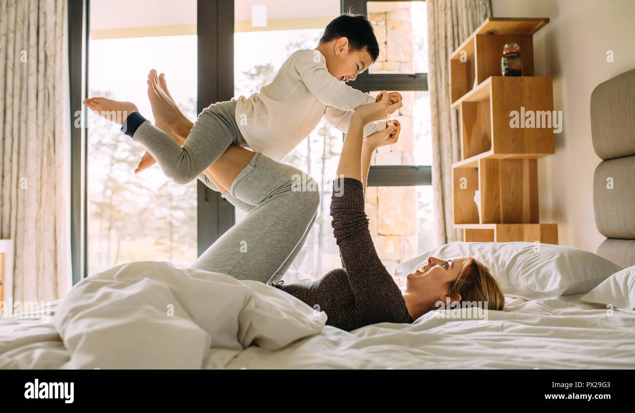 Side view of happy young woman playing with little kid on the bed. Woman lying on bed and lifting a her son with her legs. Stock Photo