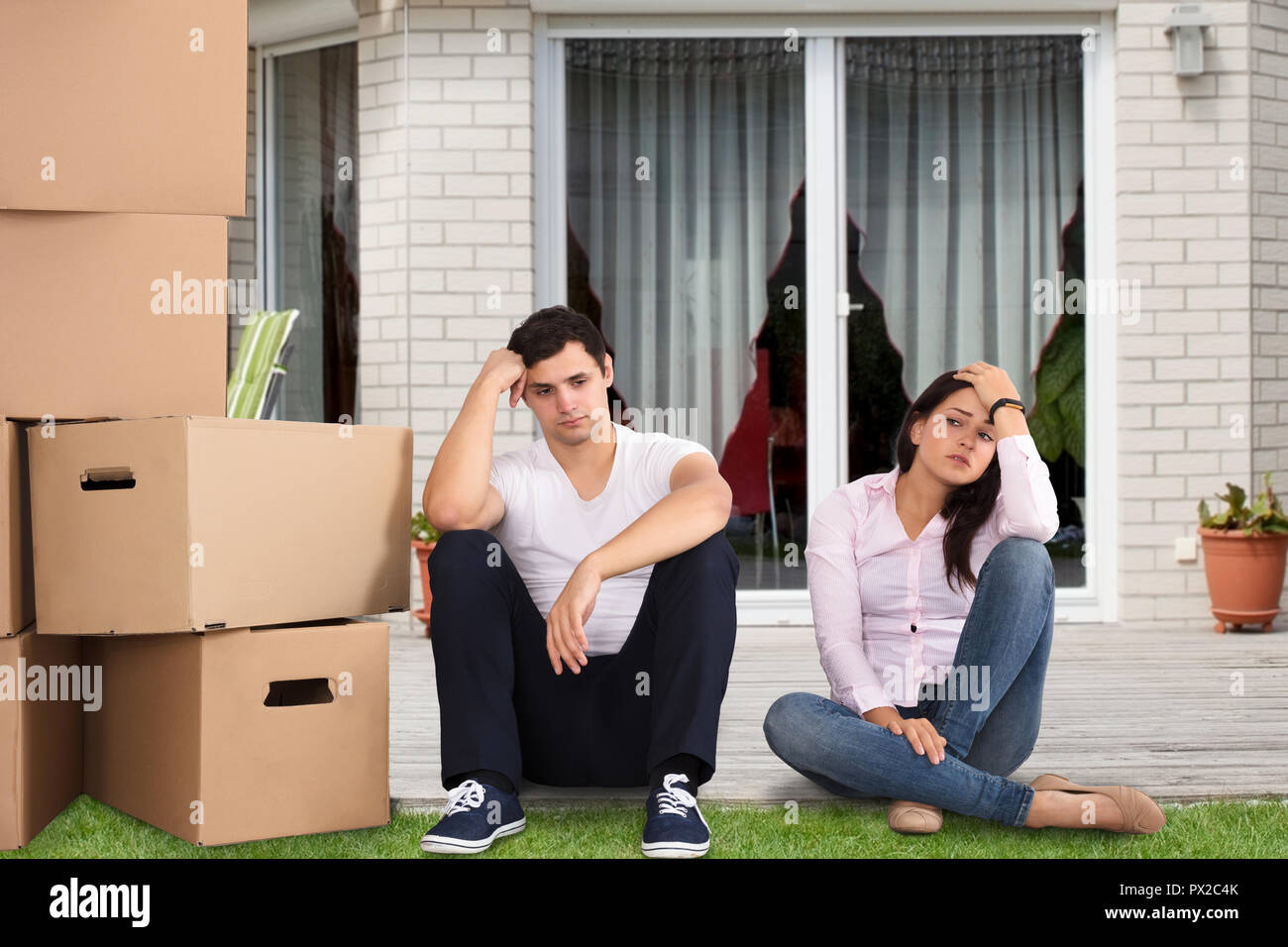 Upset Couple Sitting Near Stacked Cardboard Boxes In Front Of Their House Stock Photo
