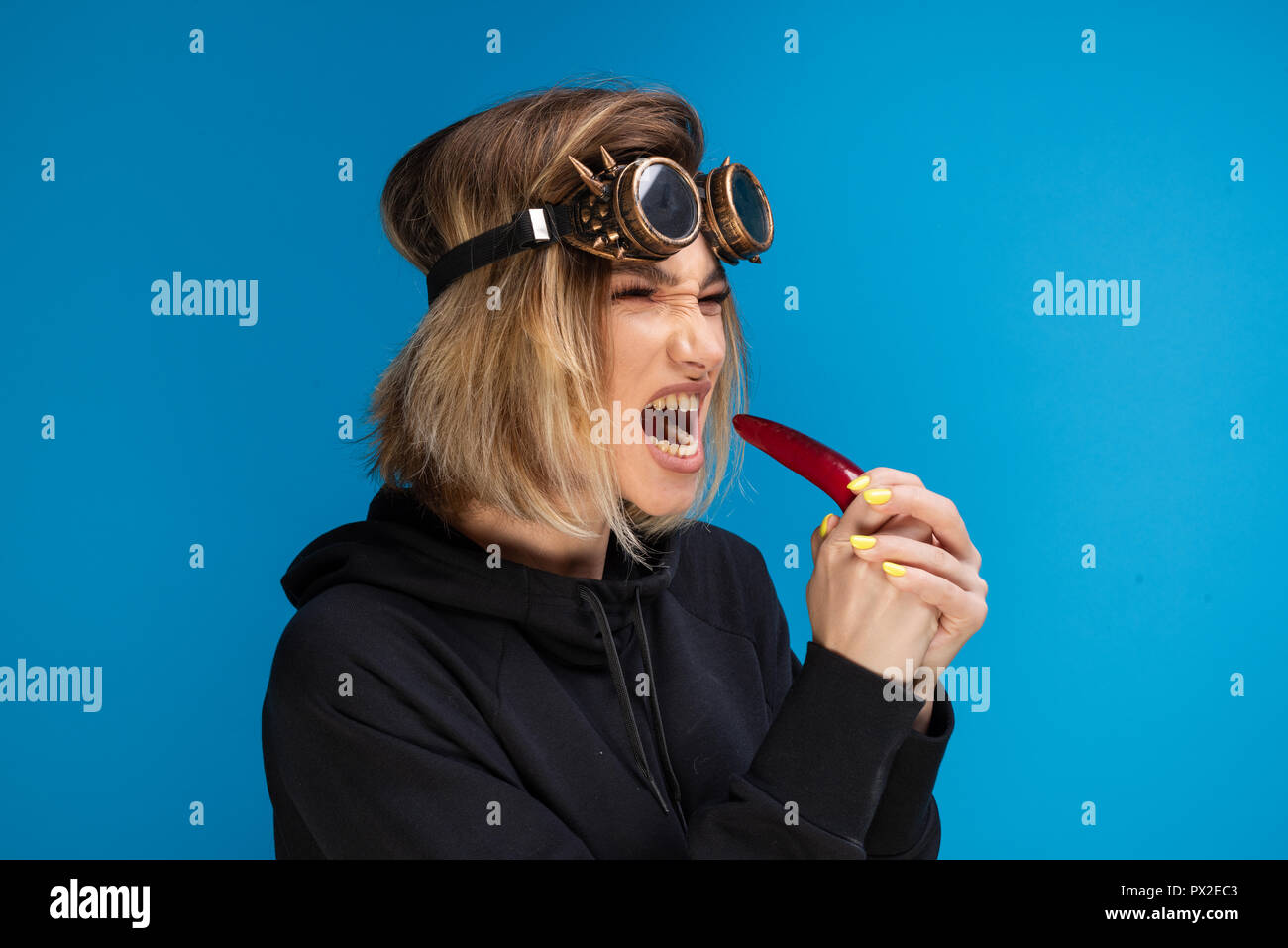 Side portrait of angry steam punk girl with dark hoodie and glasses biting a dark red chilli pepper with attitude. Dark blonde portrait shot in studio against blue background Stock Photo