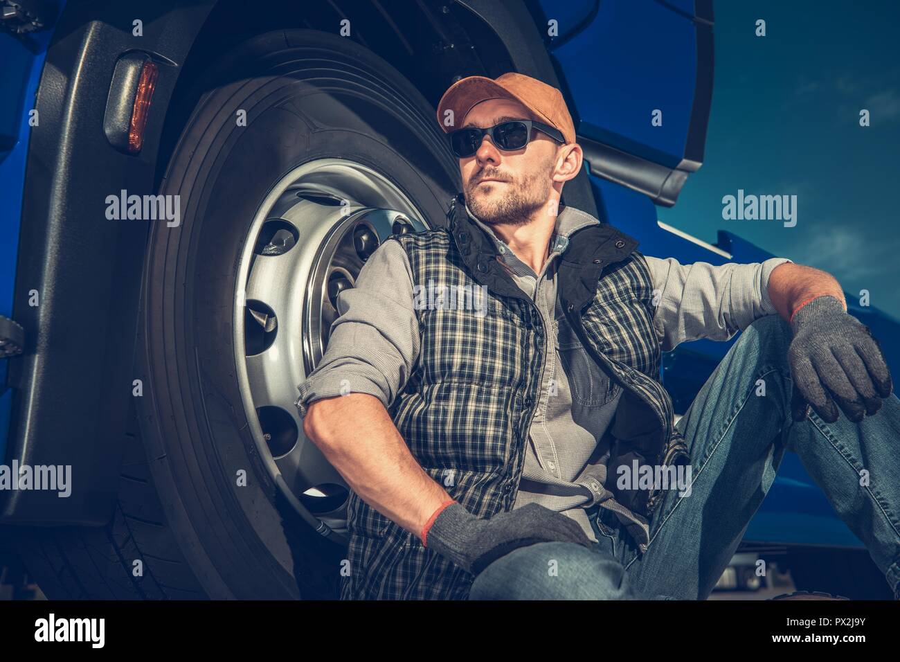 Relaxed Caucasian Truck Driver Seating on the Ground and Support His Back on the Semi Truck Wheel. Stock Photo