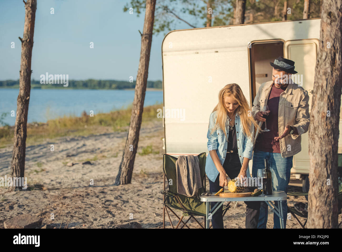 young woman cutting pineapple while man with glass of wine talking to her near trailer Stock Photo