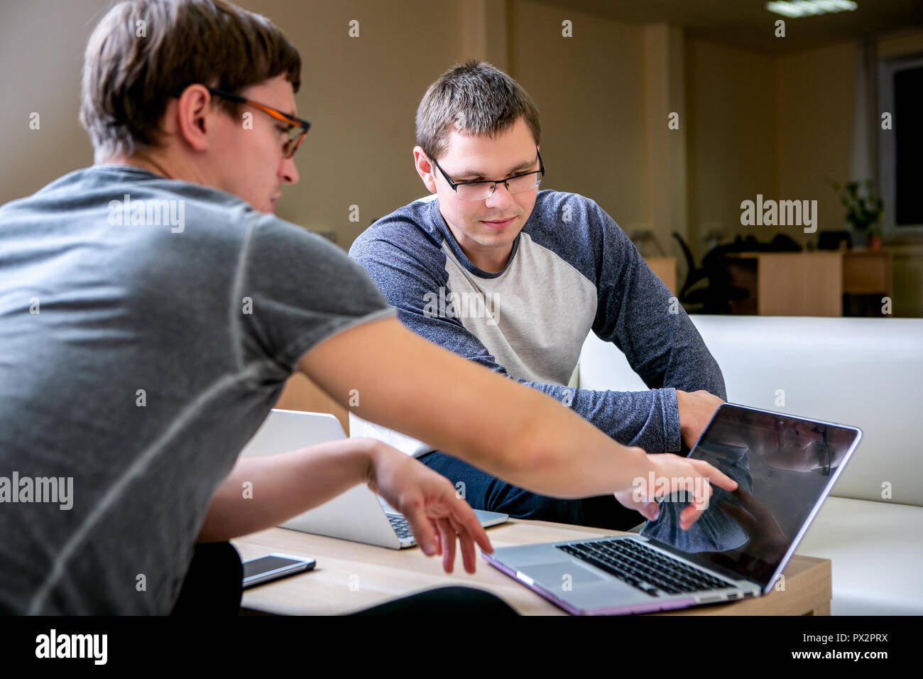 Two young people are discussing a project in the office. Sit at the table next to each other, one of them tells the other about his project and shows on the display Stock Photo