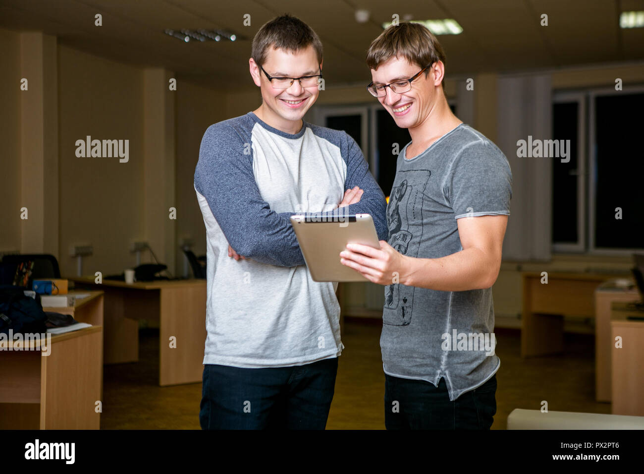 Two young people are discussing a project in the office. Standing next to each other, one of them tells the other about his project and shows on the tablet, they smile Stock Photo