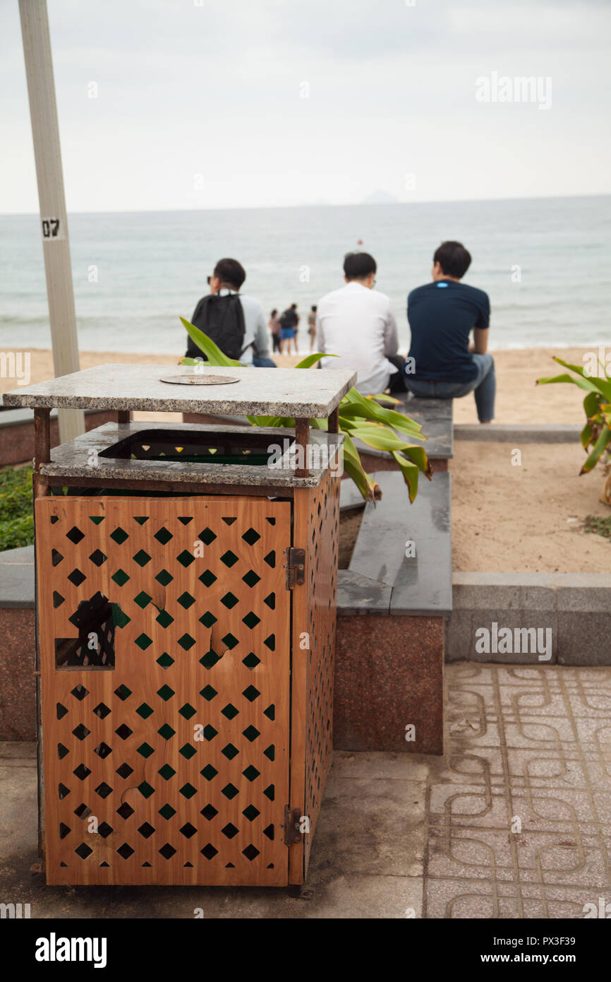 Three Young Asian man looking at the sea view. 3 people from behind sitting on a city beach near a trash bin. Focus on a trash can Stock Photo