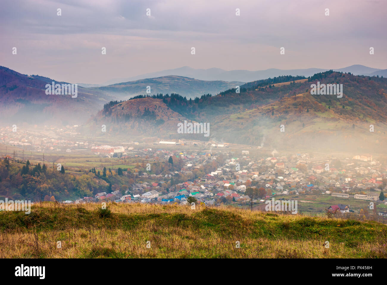 autumn countryside in mountains. small town in hazy valley. forested hills in fall colors. gloomy afternoon with overcast sky Stock Photo