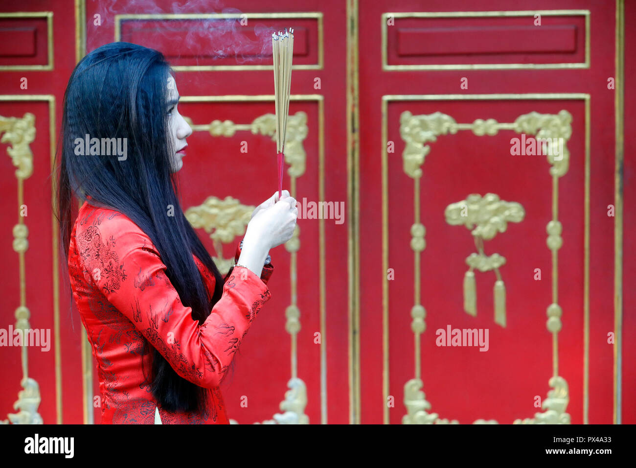 Vietnamese woman in red traditional long dress Ao Dai praying with incense sticks.  Ho Chi Minh City. Vietnam. Stock Photo