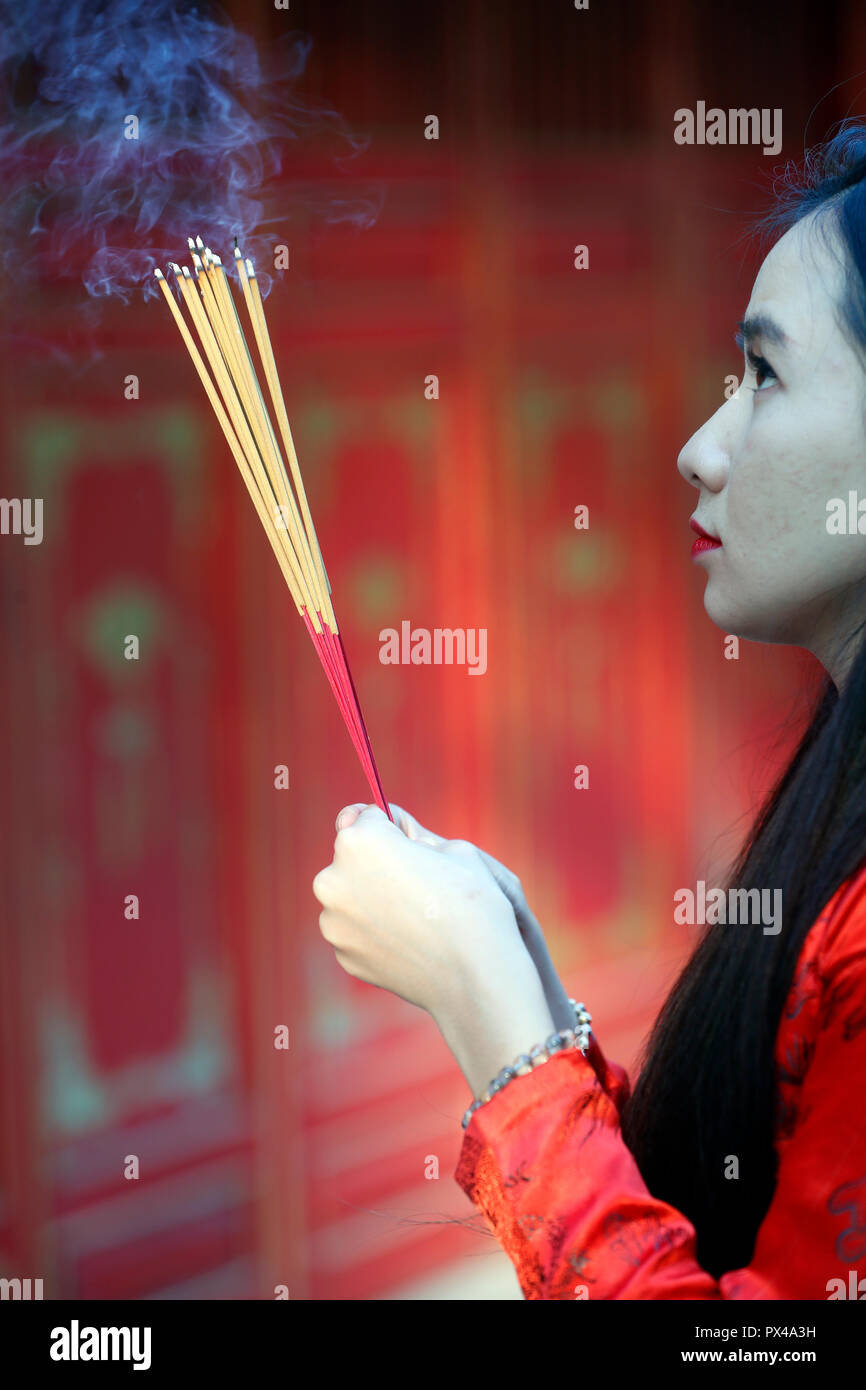 Vietnamese woman in red traditional long dress Ao Dai praying with incense sticks.  Ho Chi Minh City. Vietnam. Stock Photo