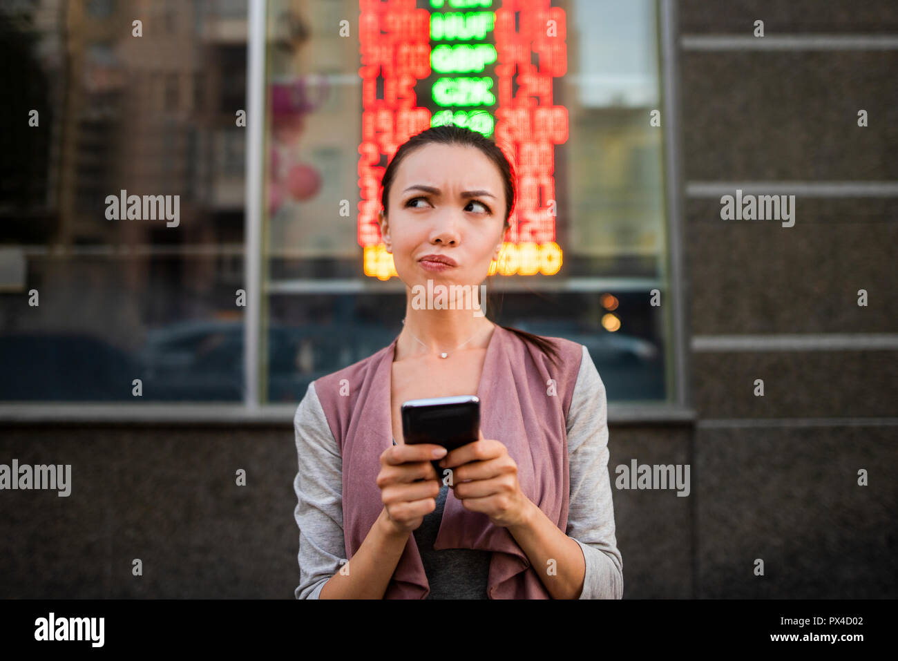 A young beautiful asian woman using an application in her smart phone to check currency exchange rates in front of an illuminated information board. Stock Photo