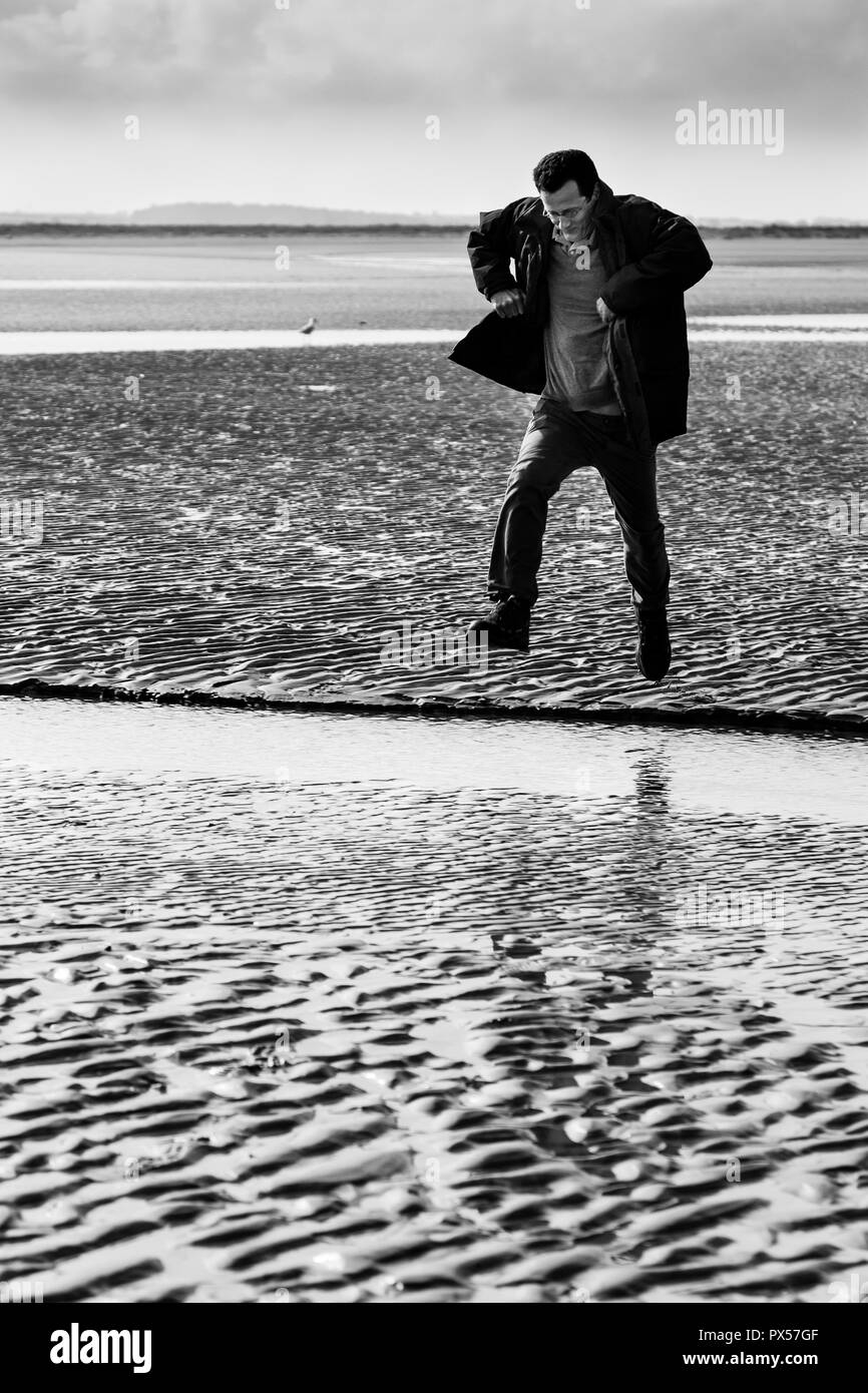 Man jumping over an inlet on the sand-flats at Brancaster, Norfolk. Stock Photo