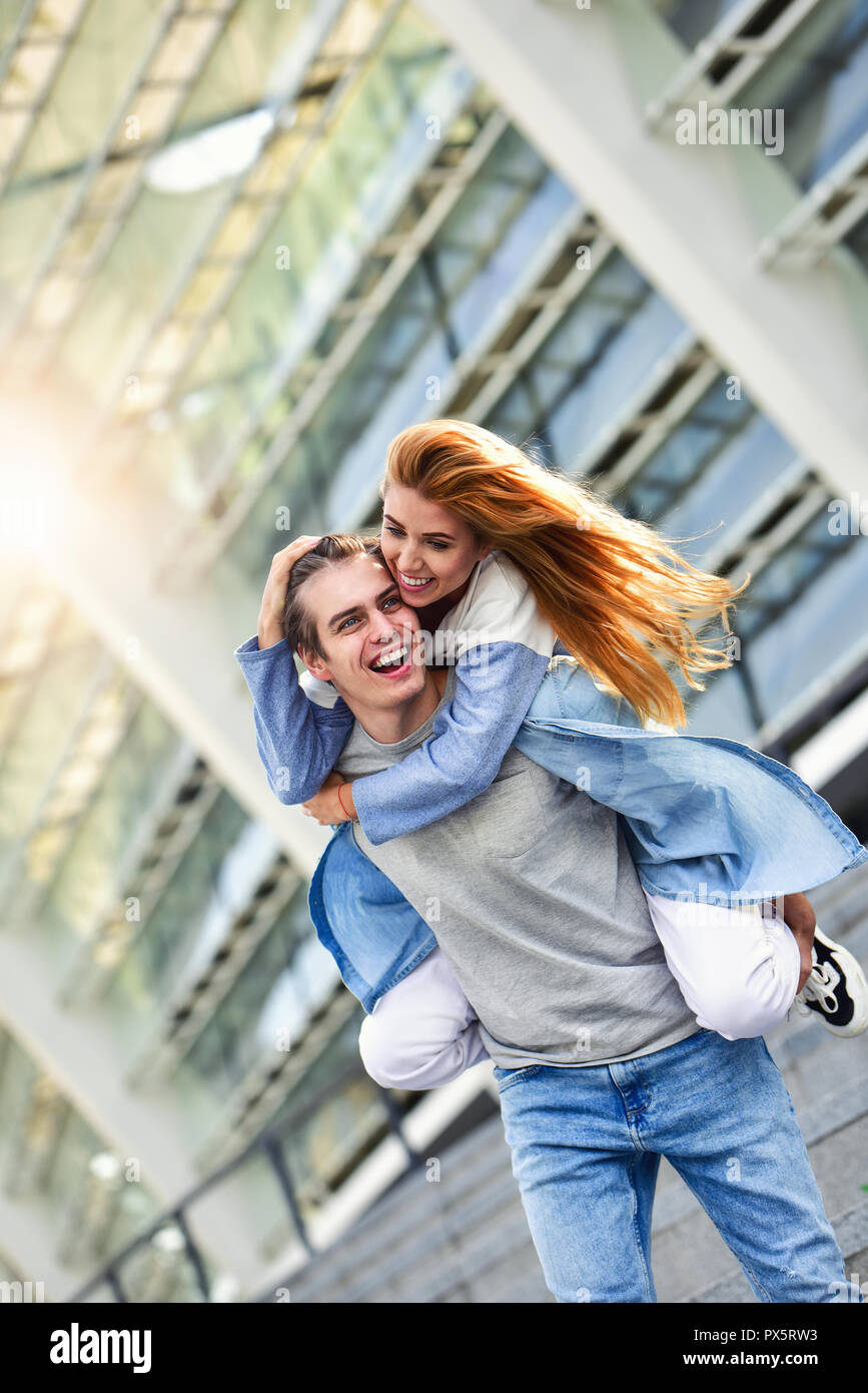 Man giving his pretty girlfriend a piggy back at street smiling at each other on a sunny day. Stock Photo