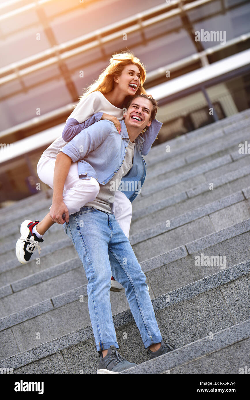 Man giving his pretty girlfriend a piggy back at street smiling at each other on a sunny day. Stock Photo
