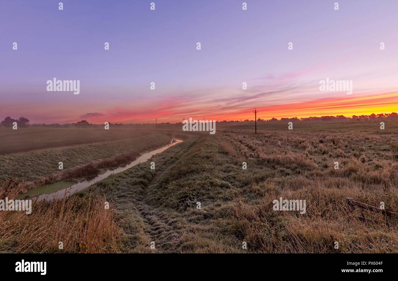 Dawn breaks across the fields with a stream running through.  A red sky is overhead. Stock Photo