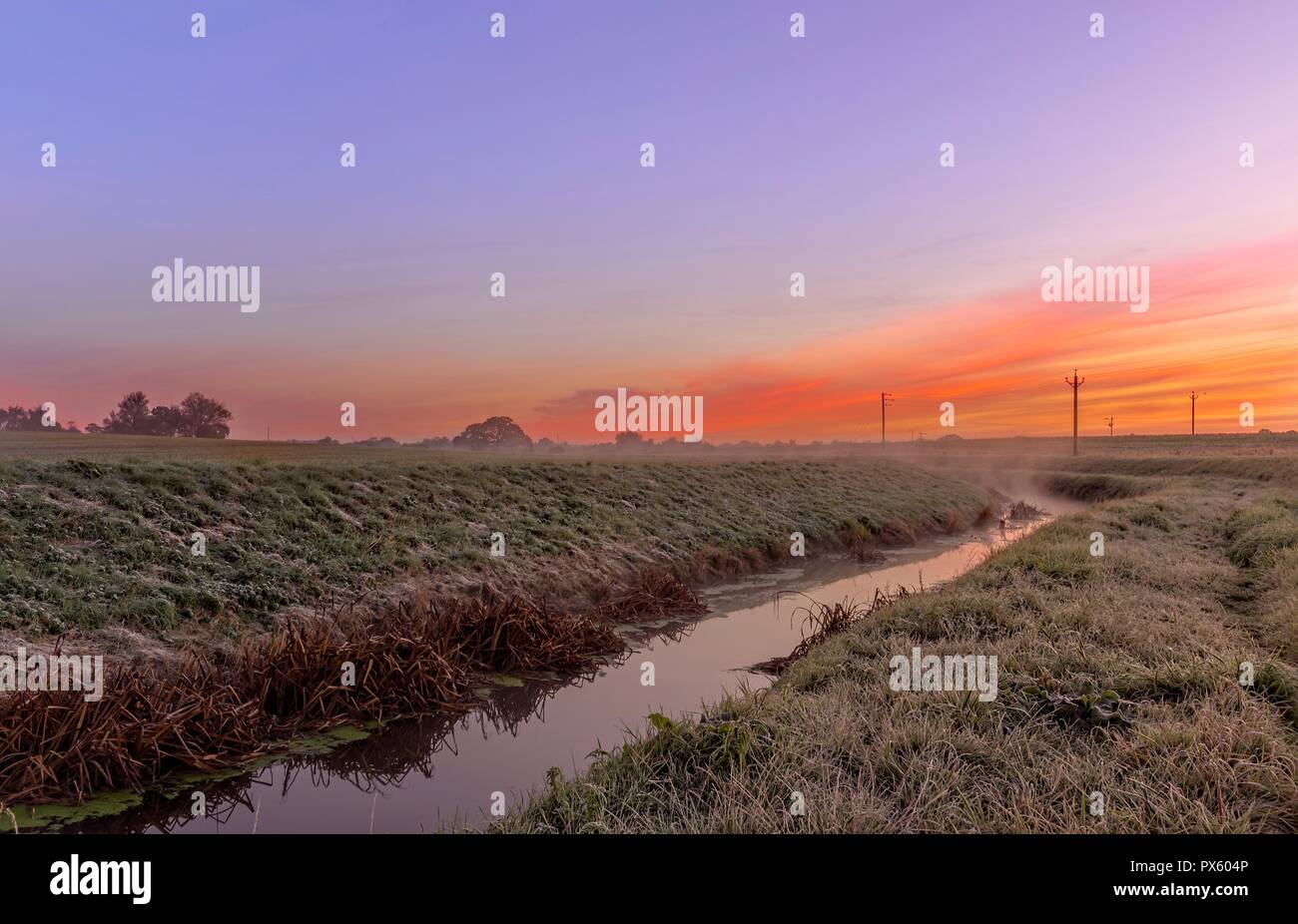 Dawn breaks across the fields with a stream running through.  A red sky is overhead. Stock Photo
