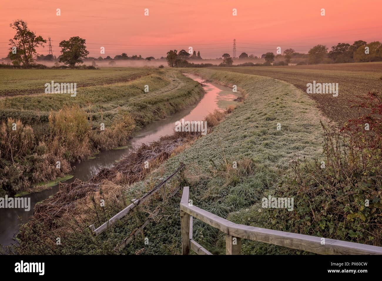 Dawn breaks across the fields with a stream running between them.  A red sky is overhead and a mist is in the distance. Stock Photo