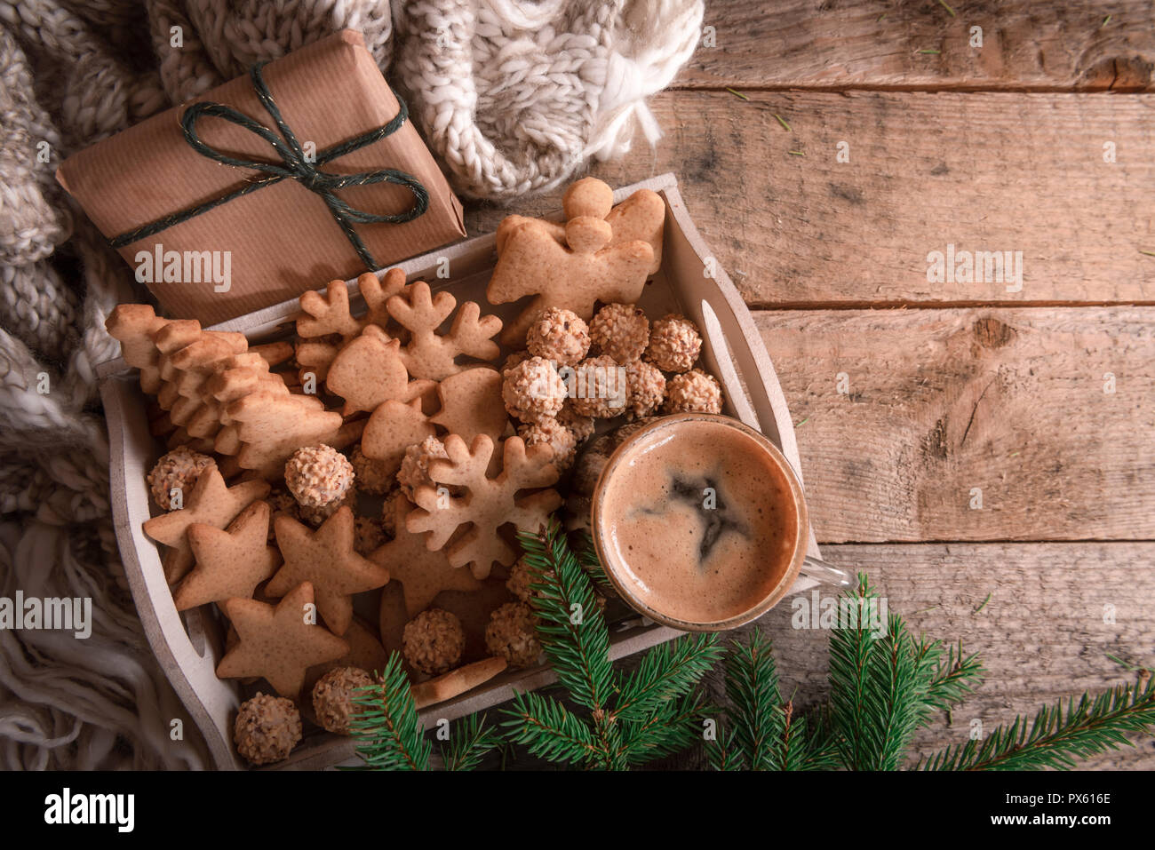 Cozy Christmas with a tray full of gingerbread, cookies and a cup of coffee, a fir twig, a gift, and a wool scarf, on a rustic table. Stock Photo