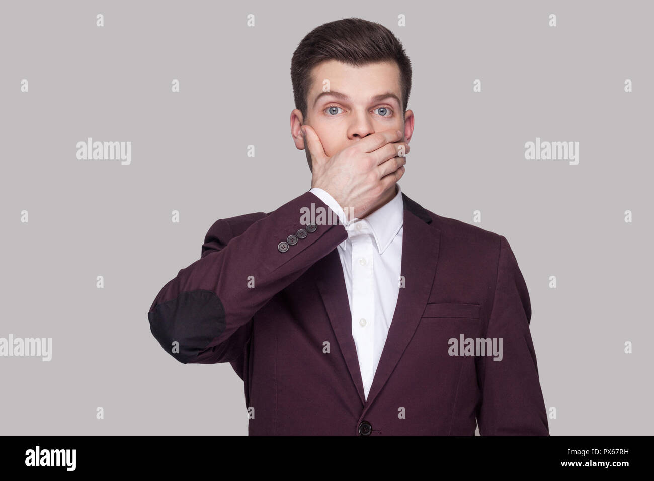 Portrait of confused handsome young man in violet suit and white shirt, standing, looking at camera with big eyes, raised arms and blocking his mouth. Stock Photo