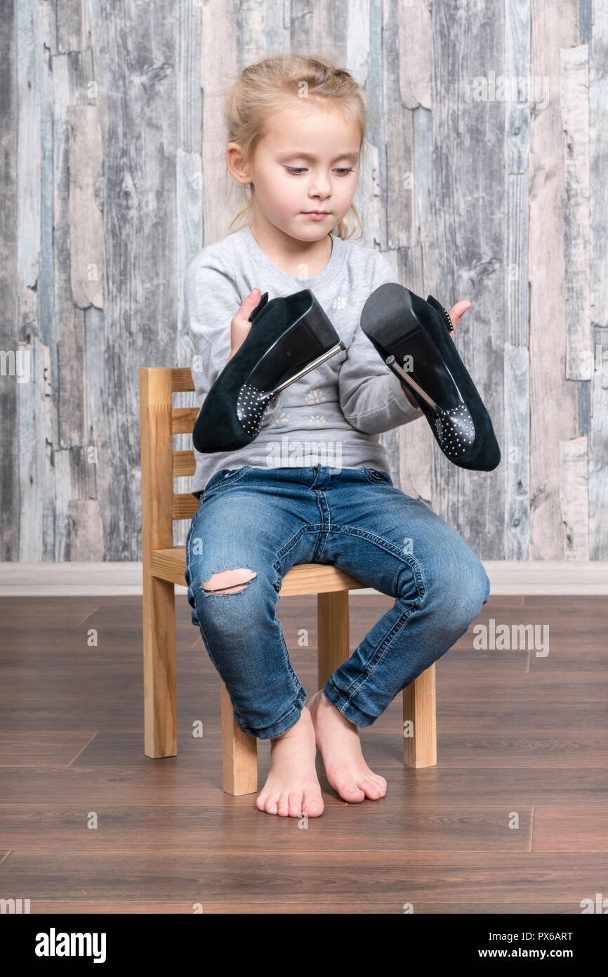little girl sits on a wooden chair and holds in her hands mother shoes with high heels, studying them Stock Photo