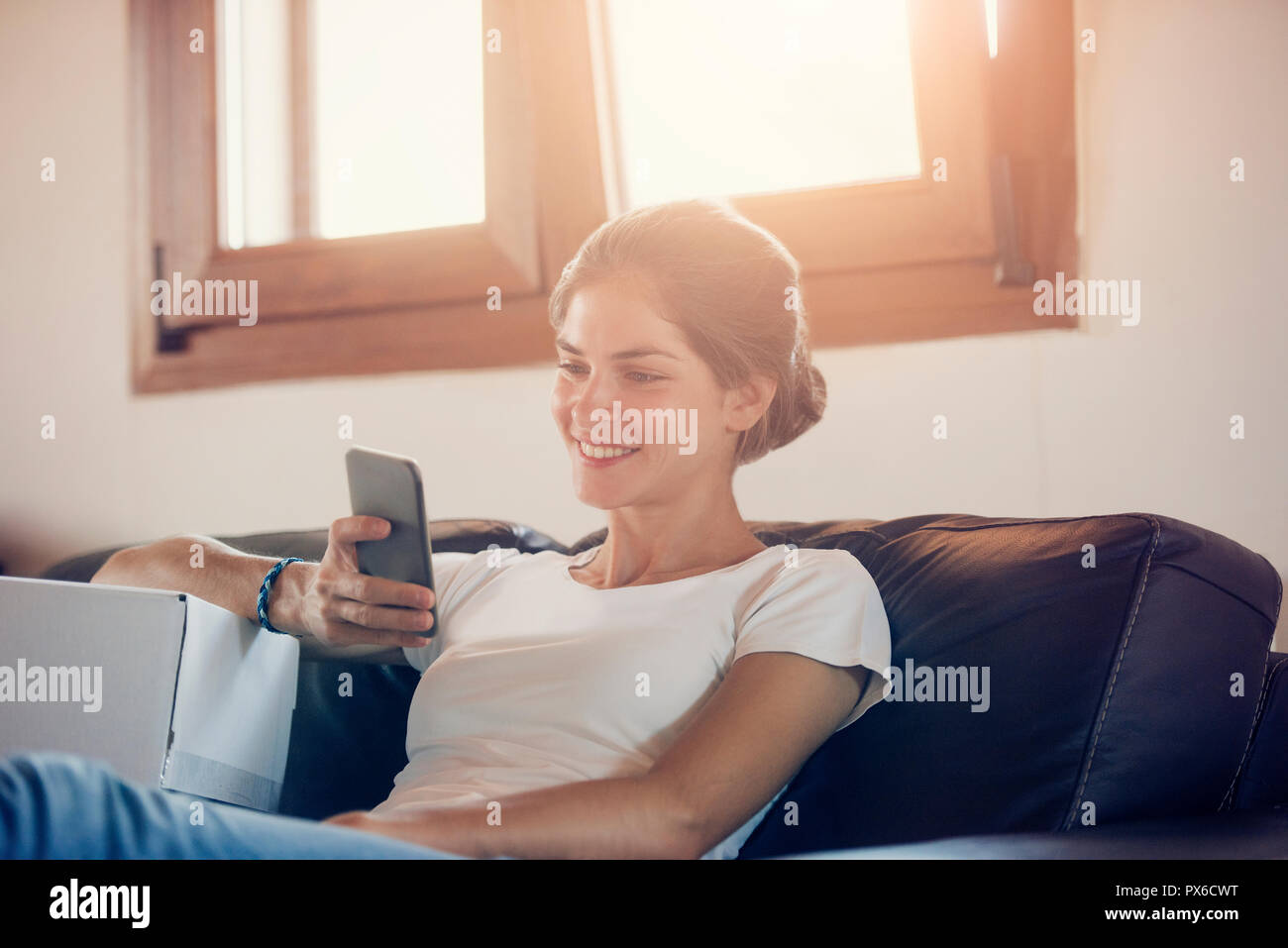 a beautiful young woman is communicating through her smart phone, next to a cardboard box. Calling for the mail delivery service. Stock Photo