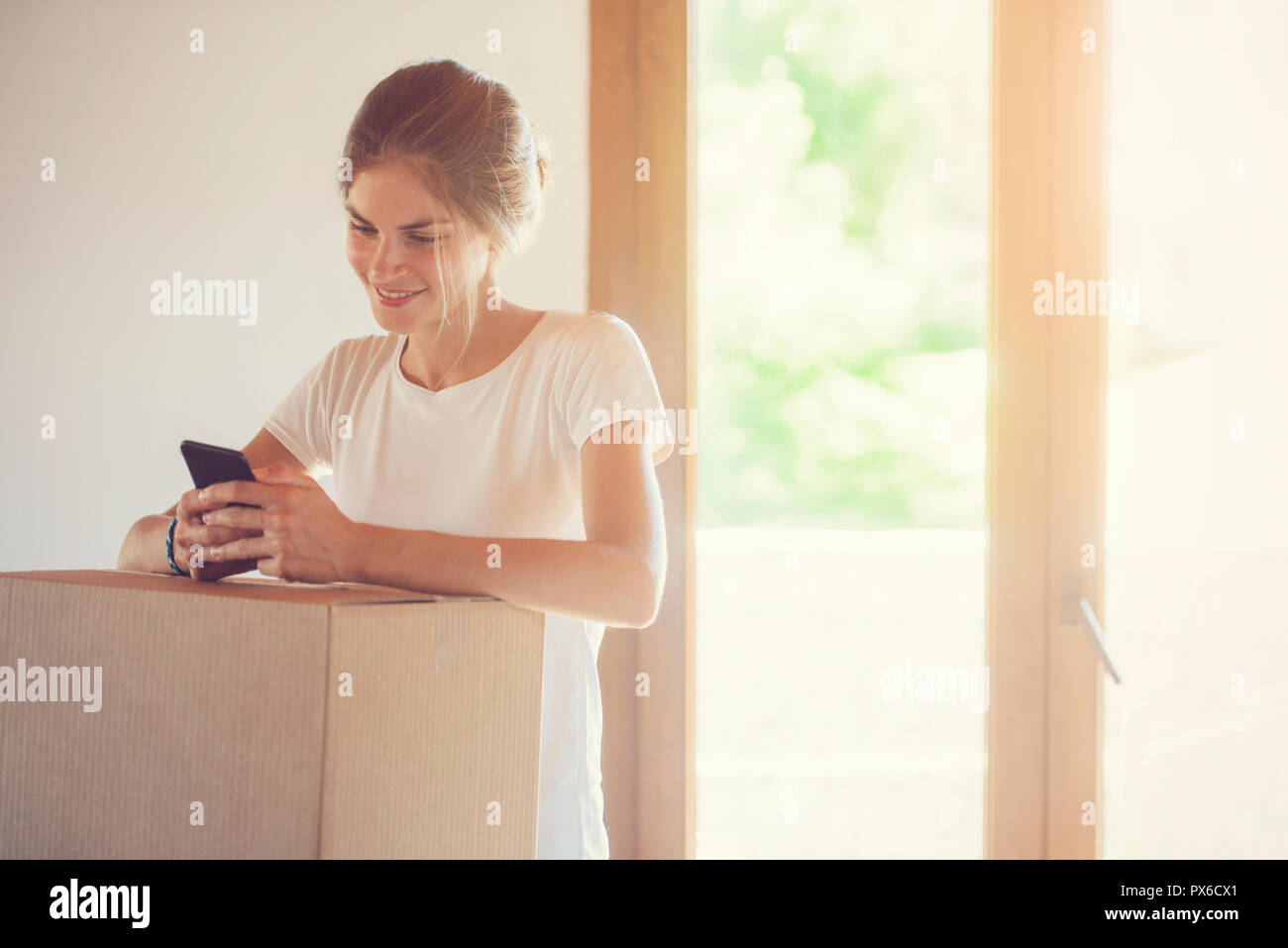 a beautiful young woman is communicating through her smart phone, next to a cardboard box. Calling for the mail delivery service. Stock Photo
