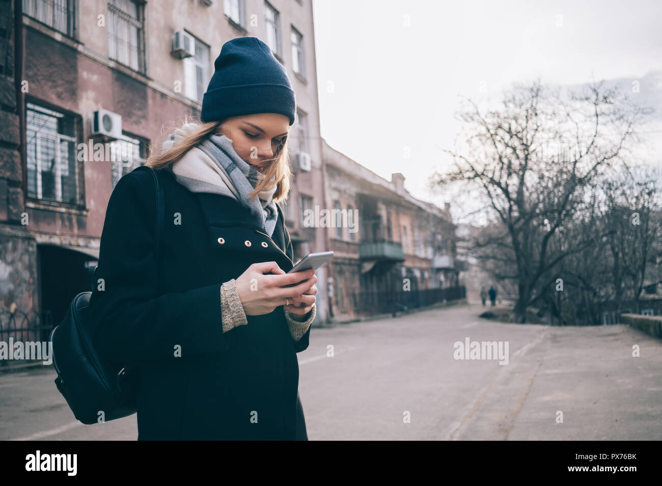Female looking at mobile device on winter street. Young caucasian woman wearing wool coat, scarf and hat using smart phone in city. Stock Photo