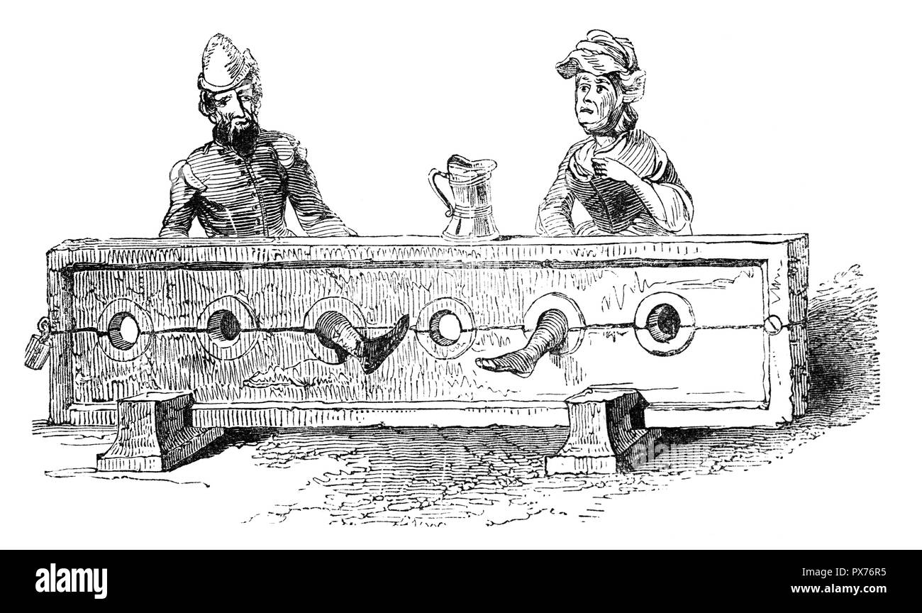 A man and woman held in Medieval Stocks,  restraining devices that were used as a form of corporal punishment and public humiliation. They consisted of of large wooden boards with hinges for restraining one's feet. The victims may be insulted, kicked, tickled, spat on, or subjected to other inhumane acts. Stock Photo