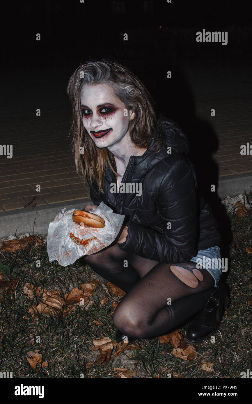 the girl in the image of a zombie with a white face in a torn t-shirt, denim shorts and black pantyhose sitting on the sidewalk and eat a Burger Stock Photo