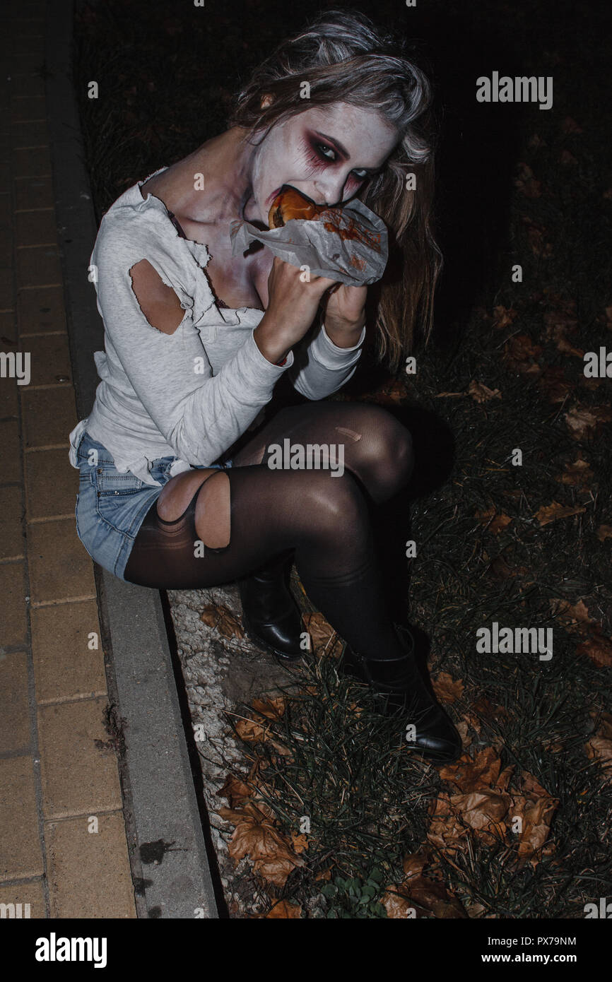 the girl in the image of a zombie with a white face in a torn t-shirt, denim shorts and black pantyhose sitting on the sidewalk and eat a Burger Stock Photo