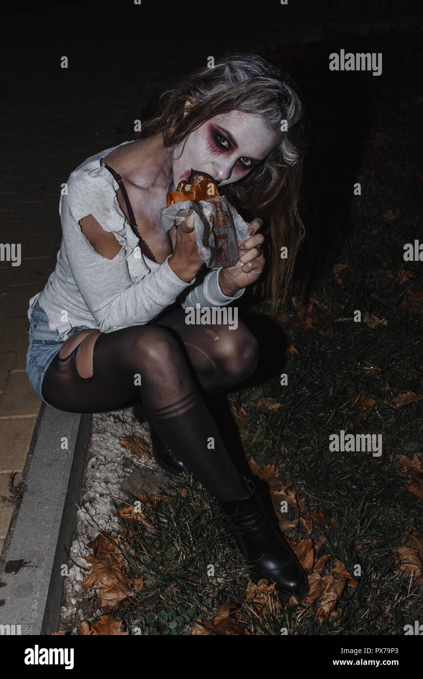 the girl in the image of a zombie with a white face in a torn t-shirt, denim shorts and black pantyhose sitting on the sidewalk and eat a Burger Stock Photo