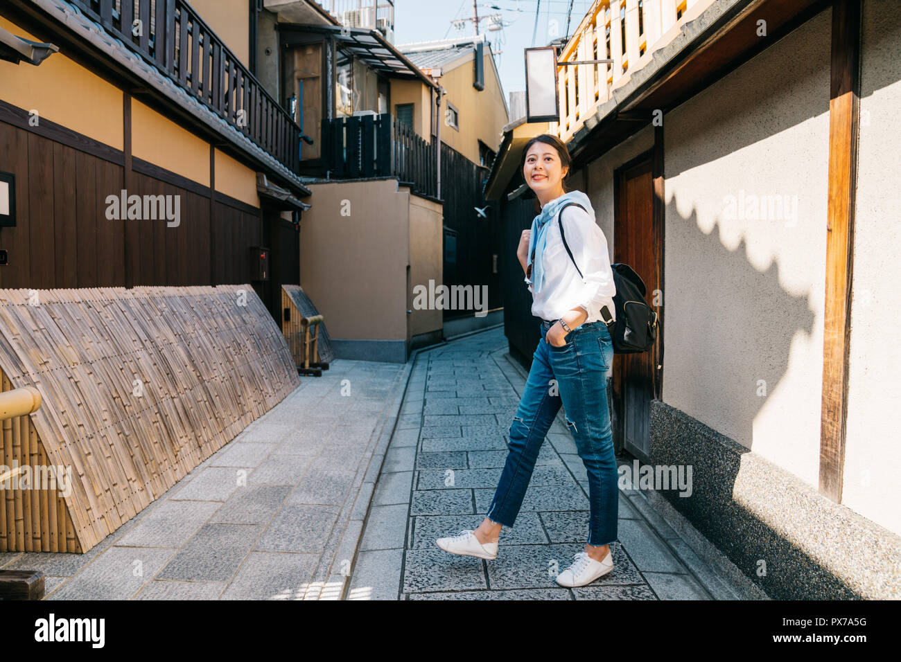 full length of a joyful woman walking in the traditional alley in Japan. backpack travel vacation young girl relaxing in path enjoying view of town is Stock Photo