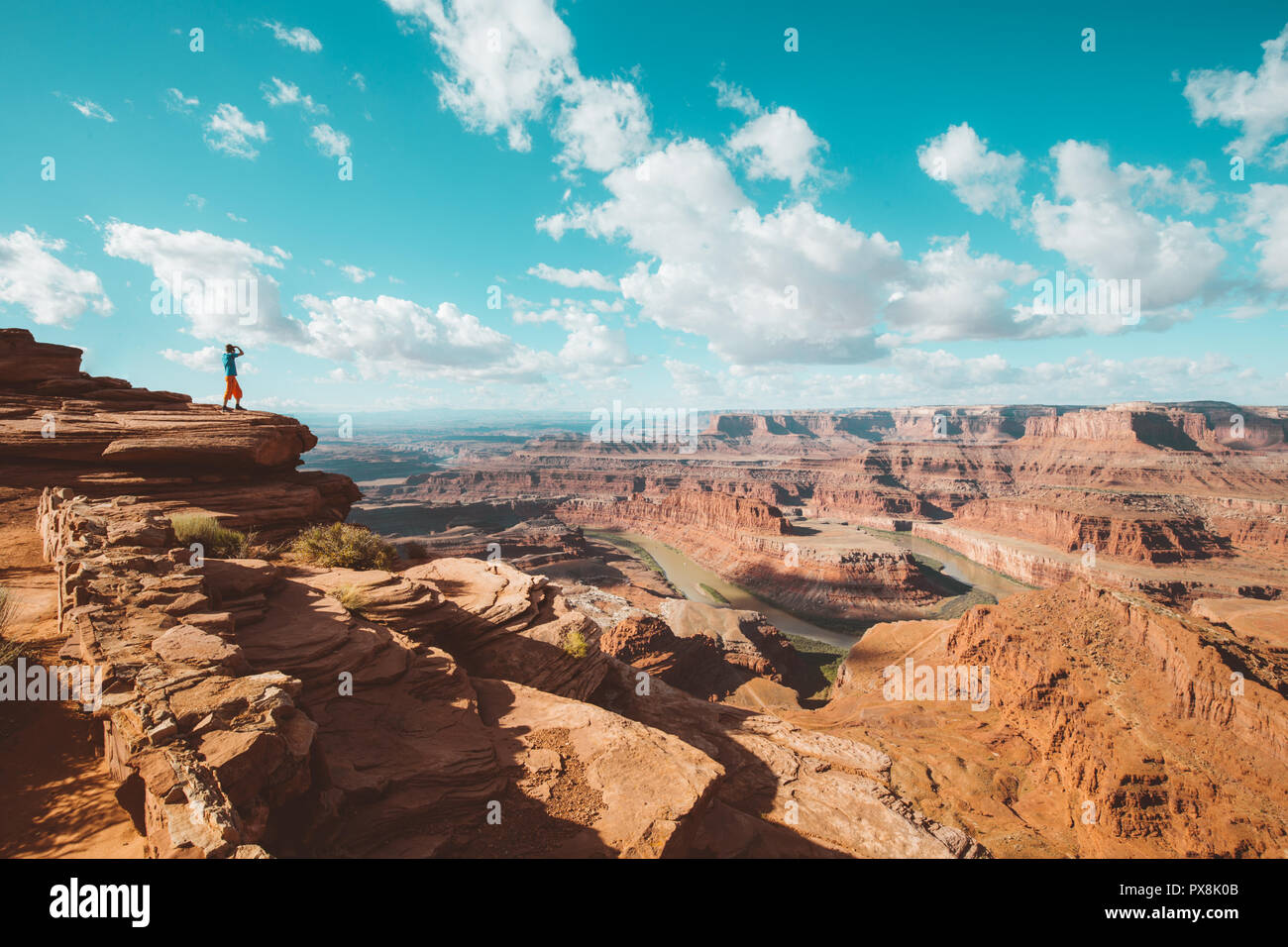 A young male hiker is standing on the edge of a cliff enjoying a dramatic view of the famous Colorado River, Dead Horse Point State Park, Utah, USA Stock Photo