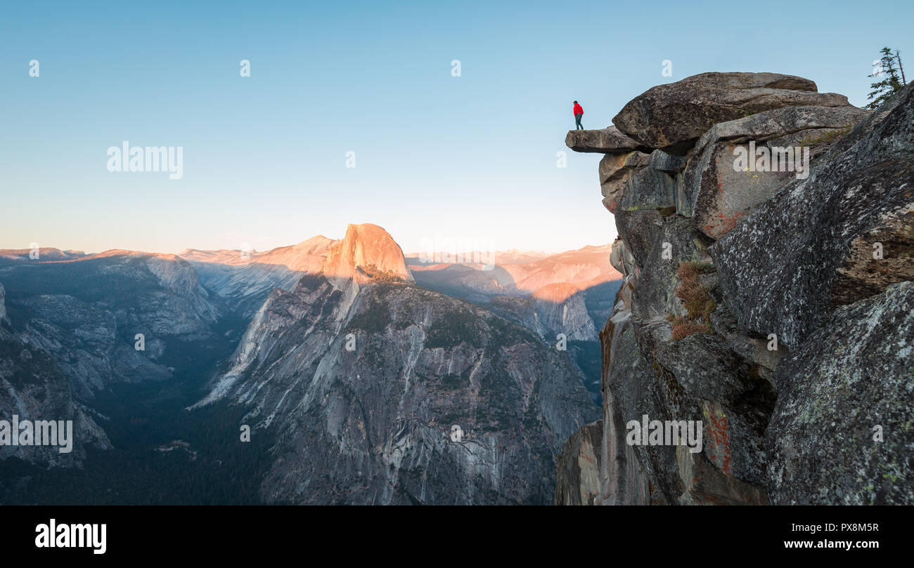 A fearless hiker is standing on an overhanging rock enjoying the view towards famous Half Dome at Glacier Point overlook in beautiful evening twilight Stock Photo