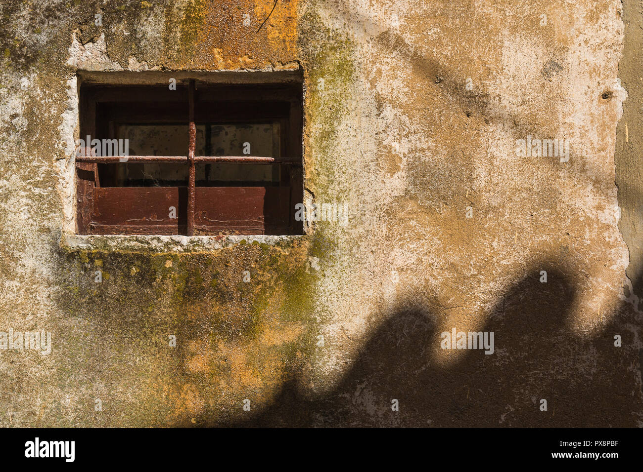 Small window with a brown frame and grating. Structured old wall, enlightened by the sunlight with a wavy shadow of another house. Vrbnik, island Krk, Stock Photo