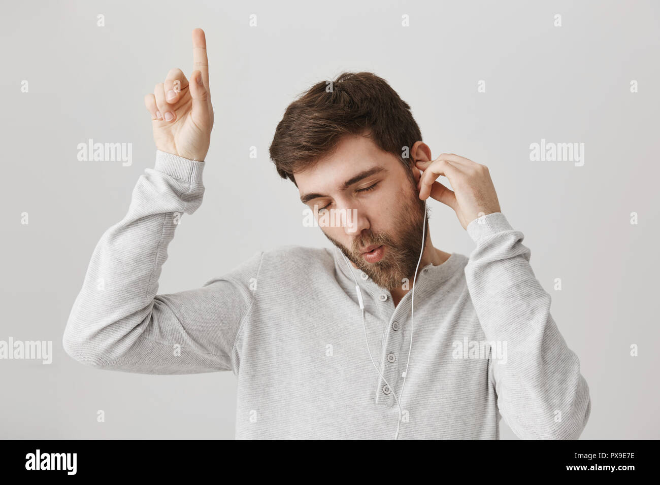Indoor shot of emotive european guy enjoying listening music via headphones, dancing with raised index finger and closed eyes over gray background. Meloman enjoying new track Stock Photo