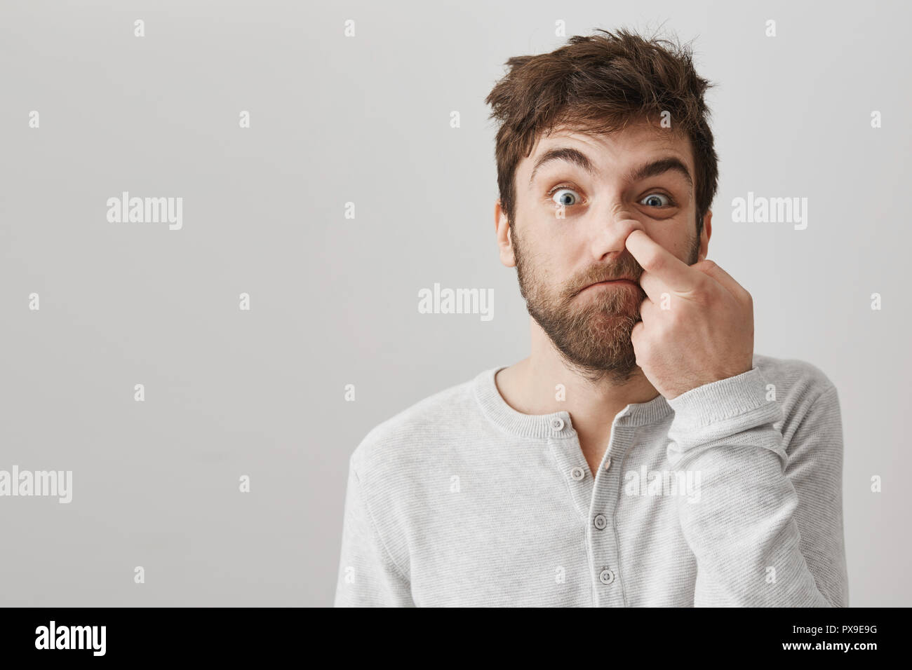 Funny bearded guy with messy hair and bad habits, staring at camera with stupid face, picking in nose with index finger and standing over gray background. Mature geek does not know about hygiene Stock Photo